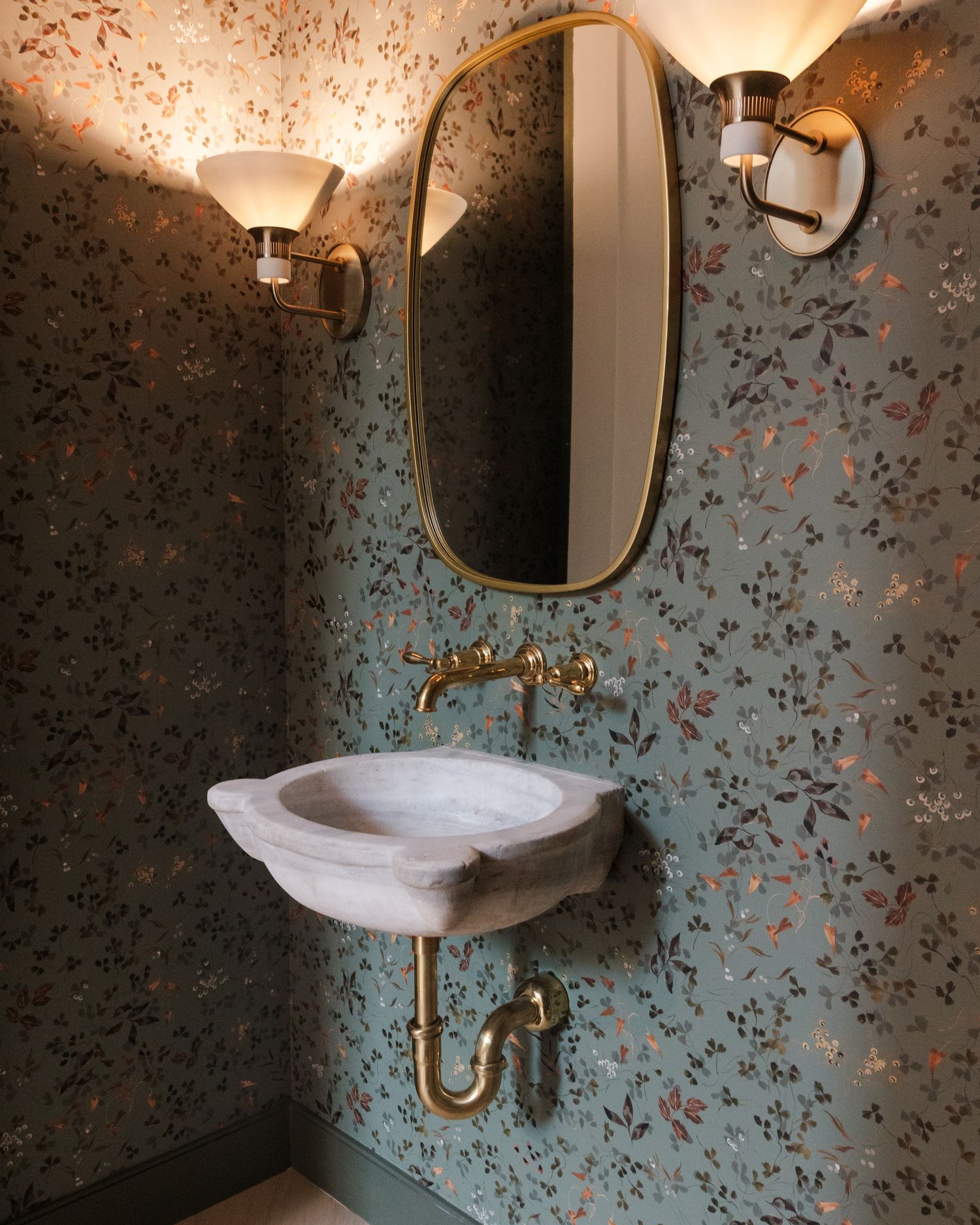 Powder bath with floral wallpaper, brass fixtures, and a wall-mounted stone sink